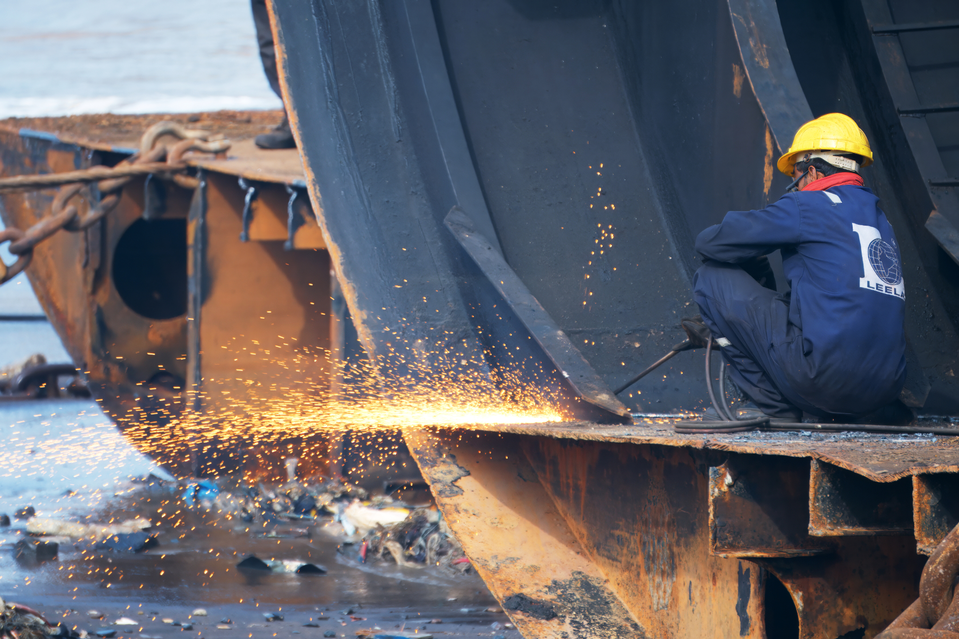 Amidst Alang's industrial symphony, a lone worker, clad in safety gear, wields a gas cutter against a steel behemoth's hull. Each spark tells a tale of grueling effort and raw heat in the heart of shipbreaking. This image captures the labor-intensive process of dismantling ships, highlighting the worker's precision and resilience. The backdrop of rusted metal and heavy machinery underscores the transformation of maritime giants into reusable resources, emphasizing the importance of safety and teamwork in this hazardous environment.